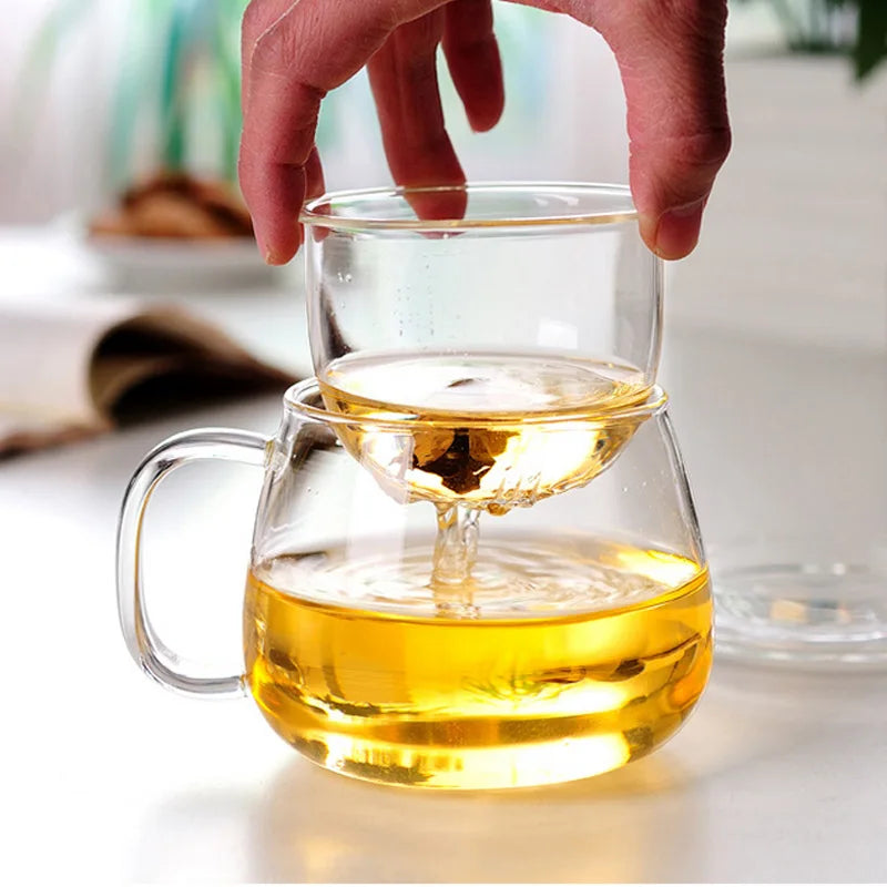 Clear glass teapot with a smaller glass cup filled with tea, held by a hand on a blurred background.