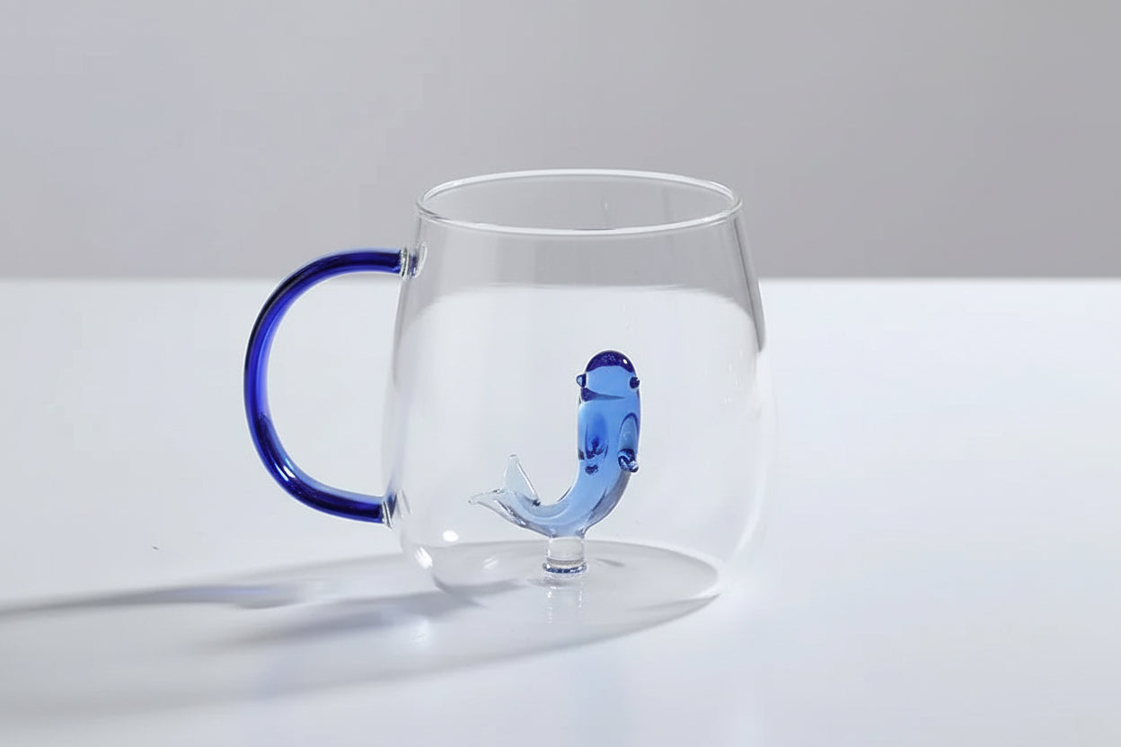 Clear glass mug with blue handle and dolphin design on a white surface