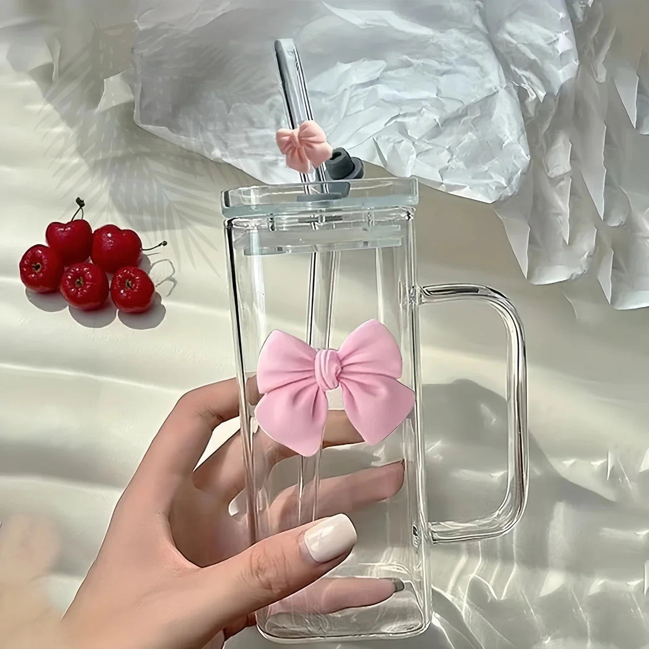 Clear mug with pink bow and handle, held by a hand, with cherries in the background.