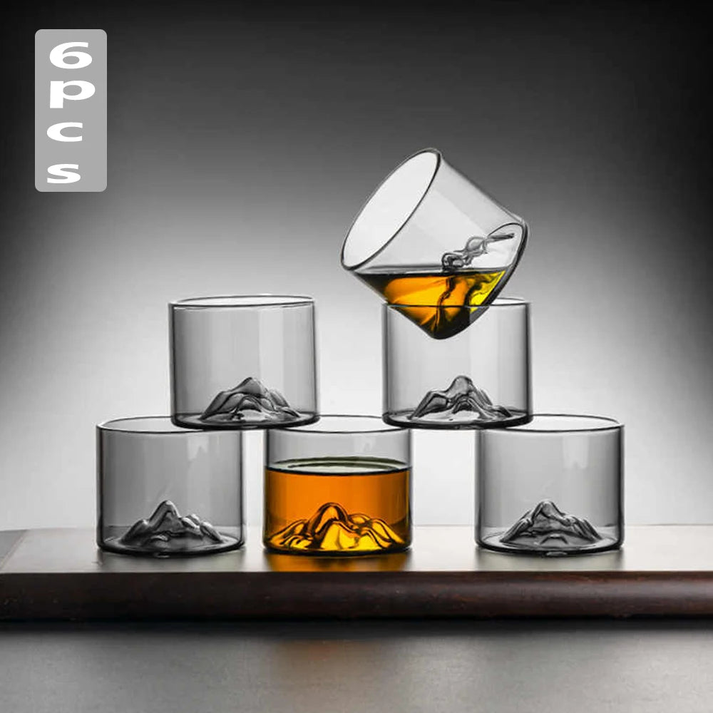Set of six clear glass cups with mountain designs, filled with amber liquid on a dark surface.