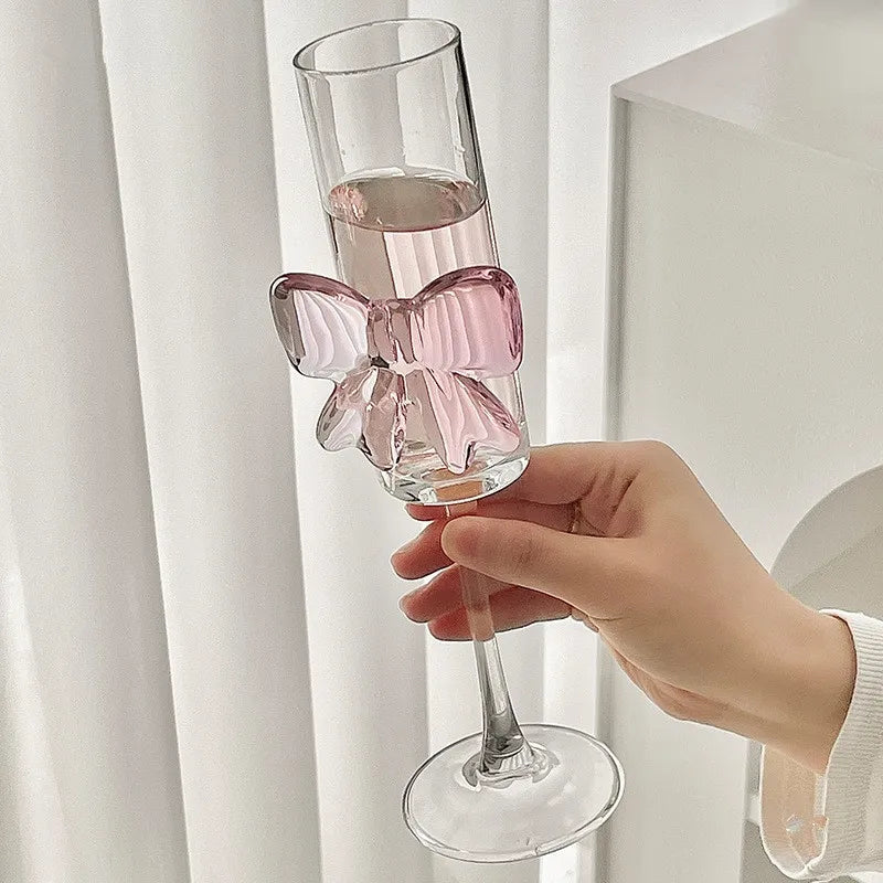 Hand holding a glass with a pink butterfly charm against a white curtain background