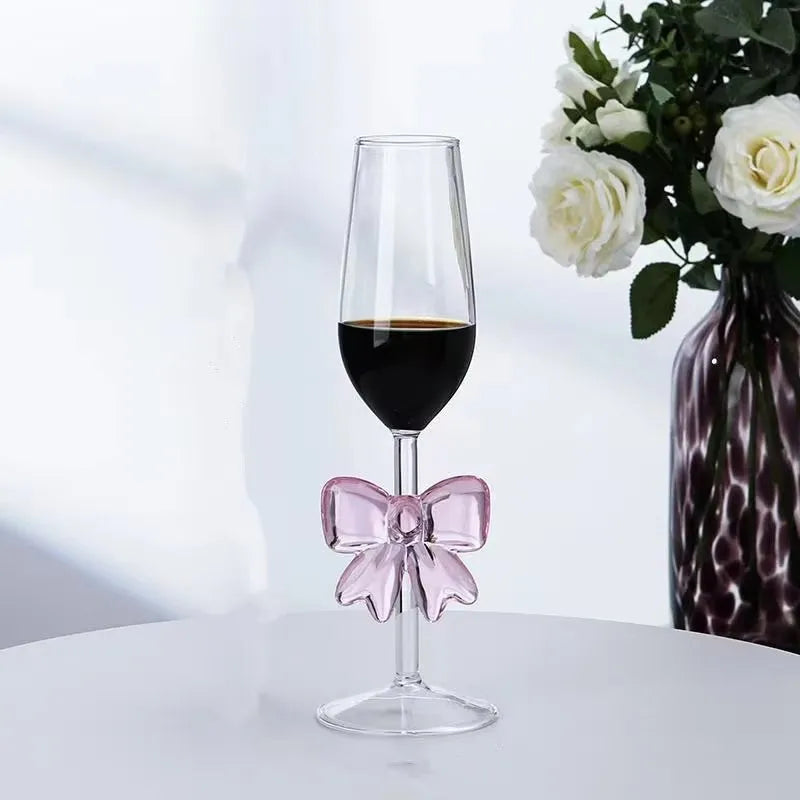Wine glass with a pink bow on a white surface, next to a vase with flowers.
