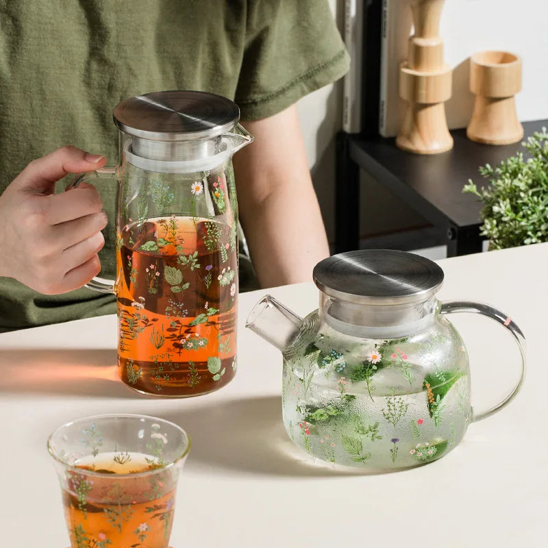 Person holding a glass teapot with floral design, next to a similar teapot and cup on a table.