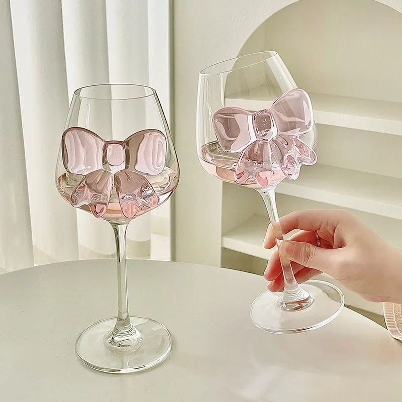 Two wine glasses with pink bow designs held by a hand on a light surface.
