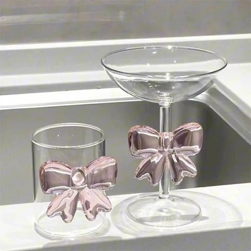 Two glass drinkware items with pink bow designs on a reflective surface.