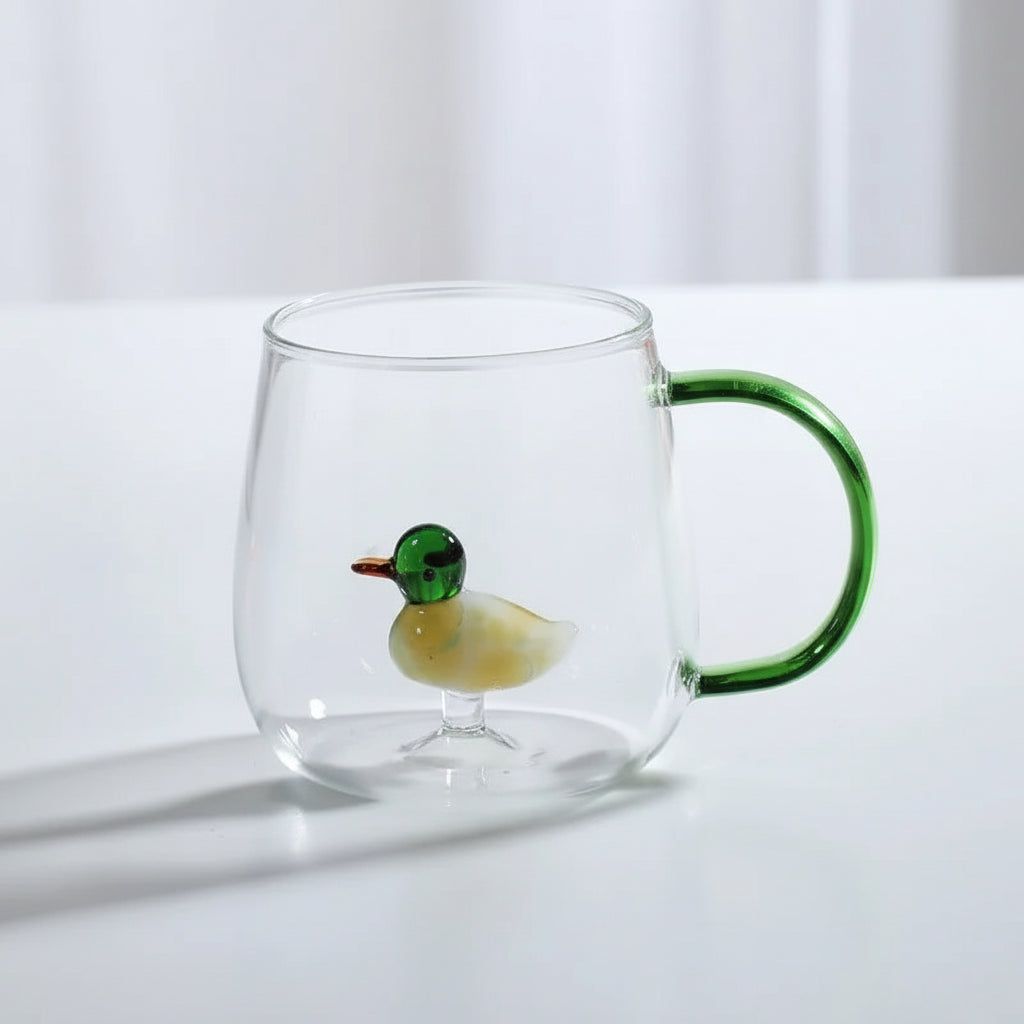 Clear glass mug with a green handle and small duck figure on a white surface.