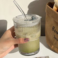Hand holding a clear glass with a green drink and straw, next to a brown paper bag with 'Bon Jour' text.